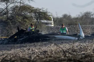 A picture taken from the crash site of the tourist plane flying over the Nazca Lines. A burnt part of the aircraft can be seen with two men stood behind it. A vehicle can be seen in the background.