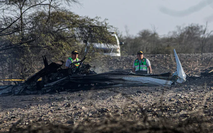 A picture taken from the crash site of the tourist plane flying over the Nazca Lines. A burnt part of the aircraft can be seen with two men stood behind it. A vehicle can be seen in the background.
