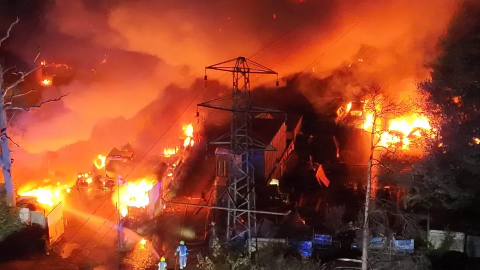 Footage from the live fire at Caldyne Business Park near Crawley during the incident, taken from above. A pylon is visible in the centre of the image, with trees half pictures each side, peering over the blaze, which is orange and yellow. Smoke billows out and around, blurring the background. Firefighters can be see on the ground attempting to control and put out the fire. 