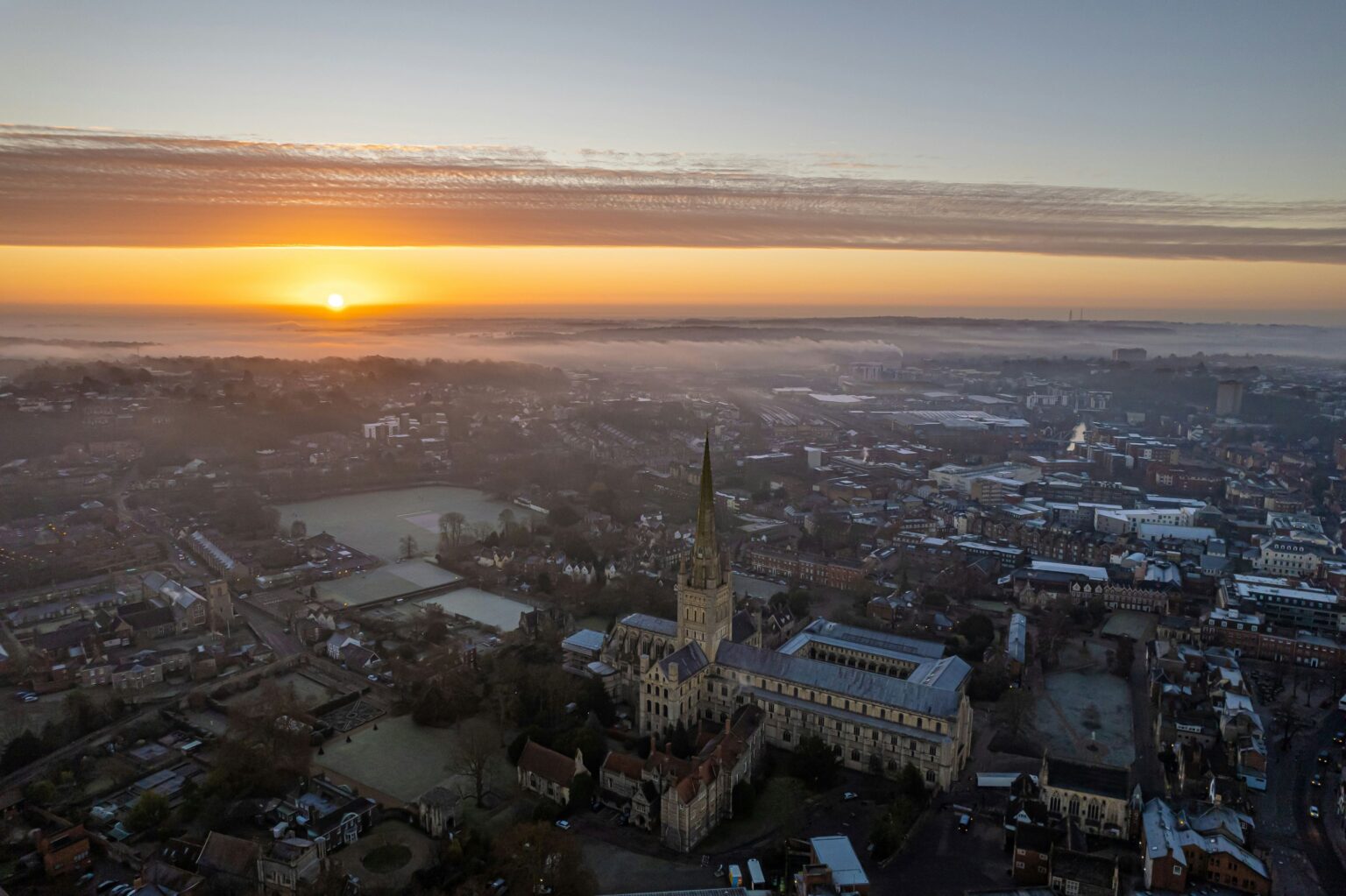 Photograph of Norwich city centre from above at sunset or sunrise, which can be seen on the horizon.