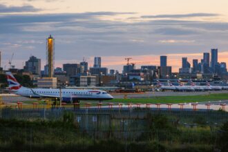 British Airways aircraft on an airport taxiway at sunset, with multiple parked planes and a city skyline in the background