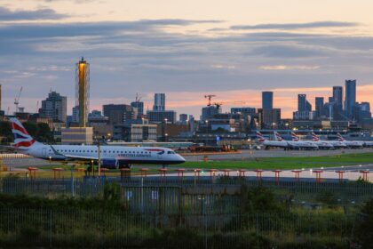 British Airways aircraft on an airport taxiway at sunset, with multiple parked planes and a city skyline in the background