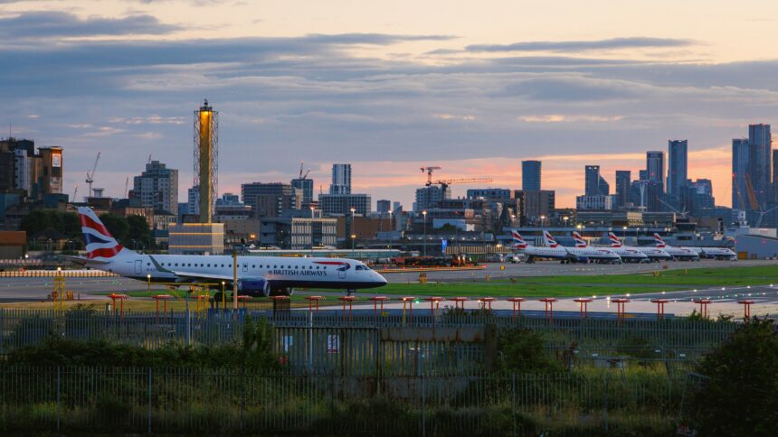 British Airways aircraft on an airport taxiway at sunset, with multiple parked planes and a city skyline in the background