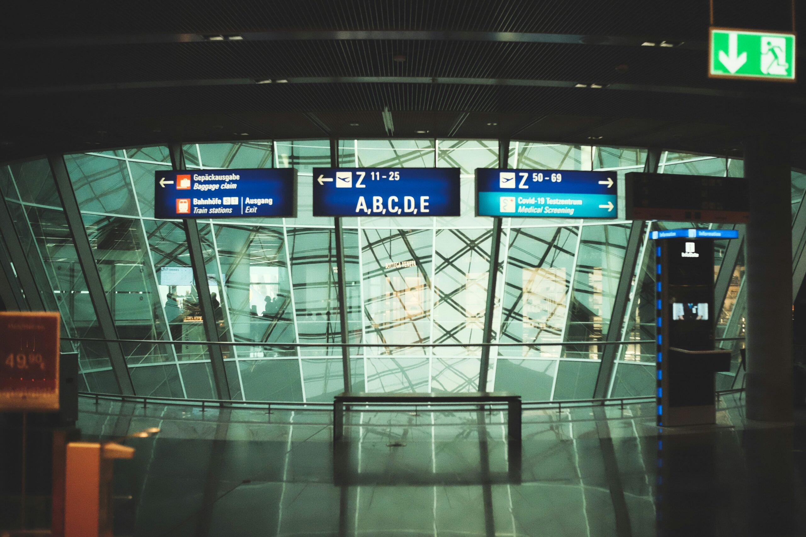 Inside of a German airport with signs pointing to different terminals. The colour theme is dark and moody, and there are multiple reflections caused by the sun shining through glass windows.