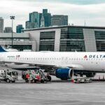 A Delta Air lines aircraft parked on tarmac with a cloudy but bright sky background and a terminal in the background.