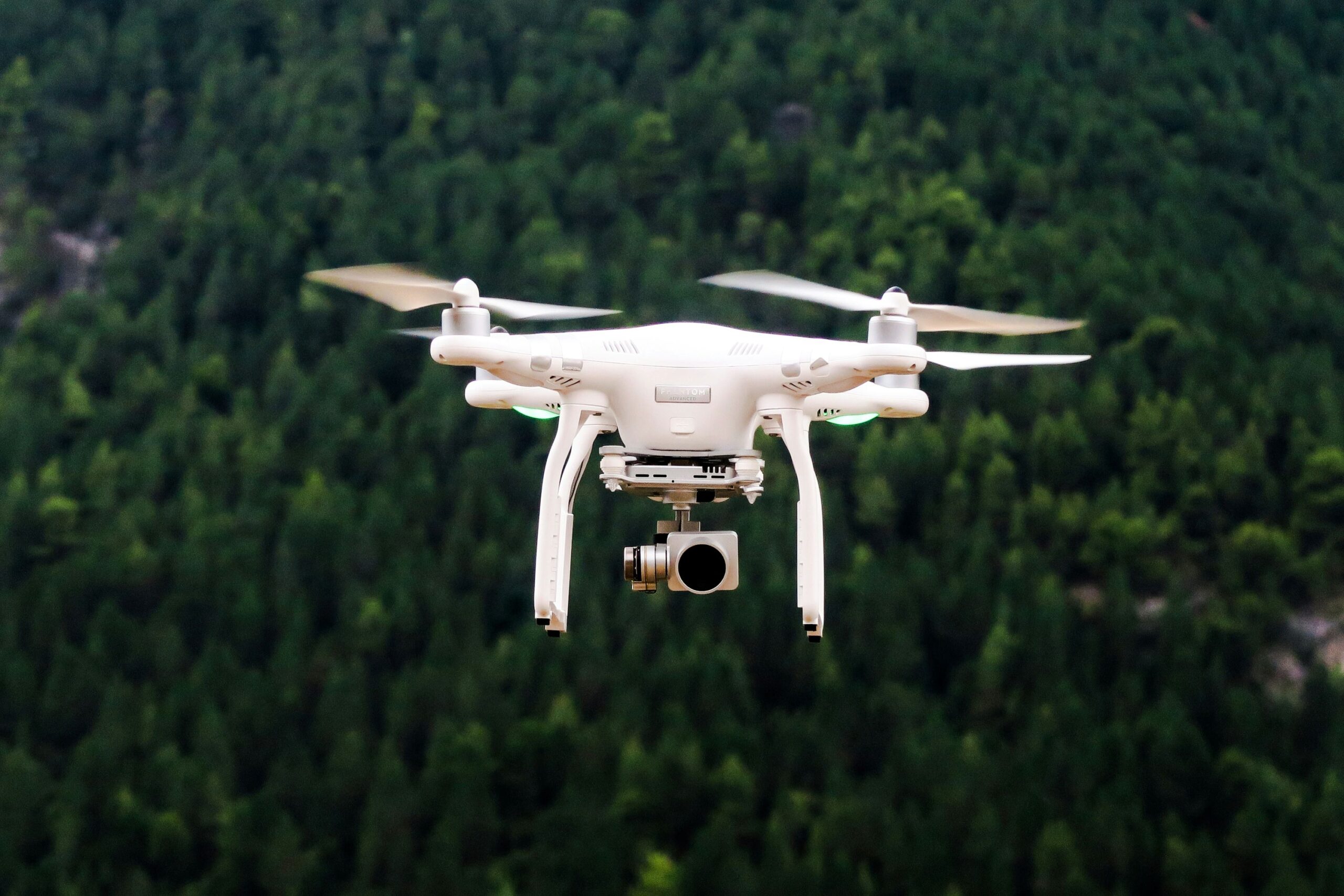 A white drone is pictured in flight. It is in the centre of the image, against a stylistically blurred background of a dense forest consisting of luscious green trees. 