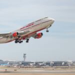 Air India double decker aircraft midair in a take off position it has a red tail and wing tips and the tarmac can be seen in the horizon with a cloudy sky