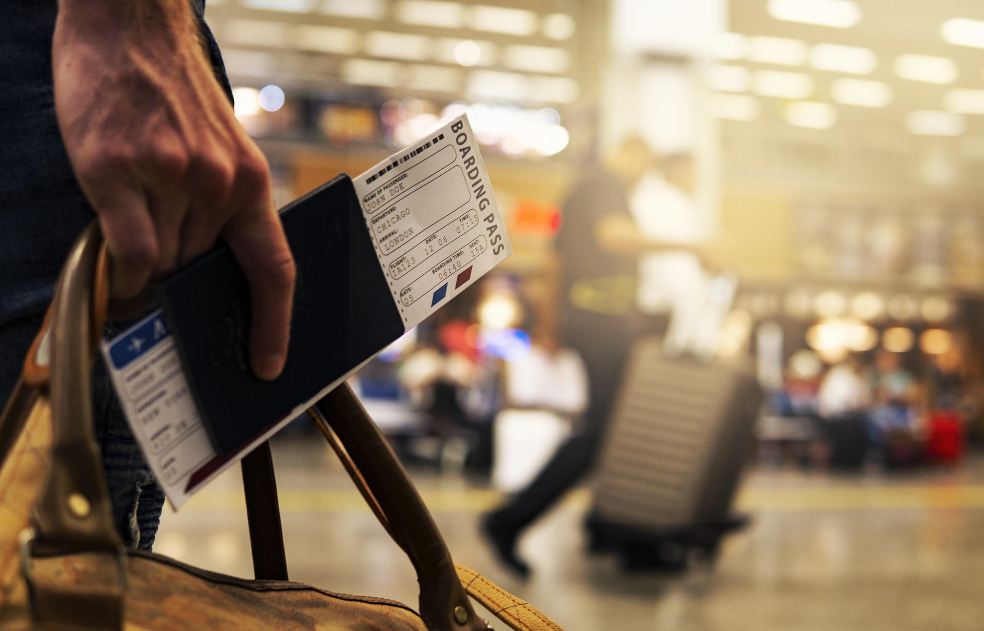 Hand holding a passport and boarding pass beside a travel bag in a busy airport terminal