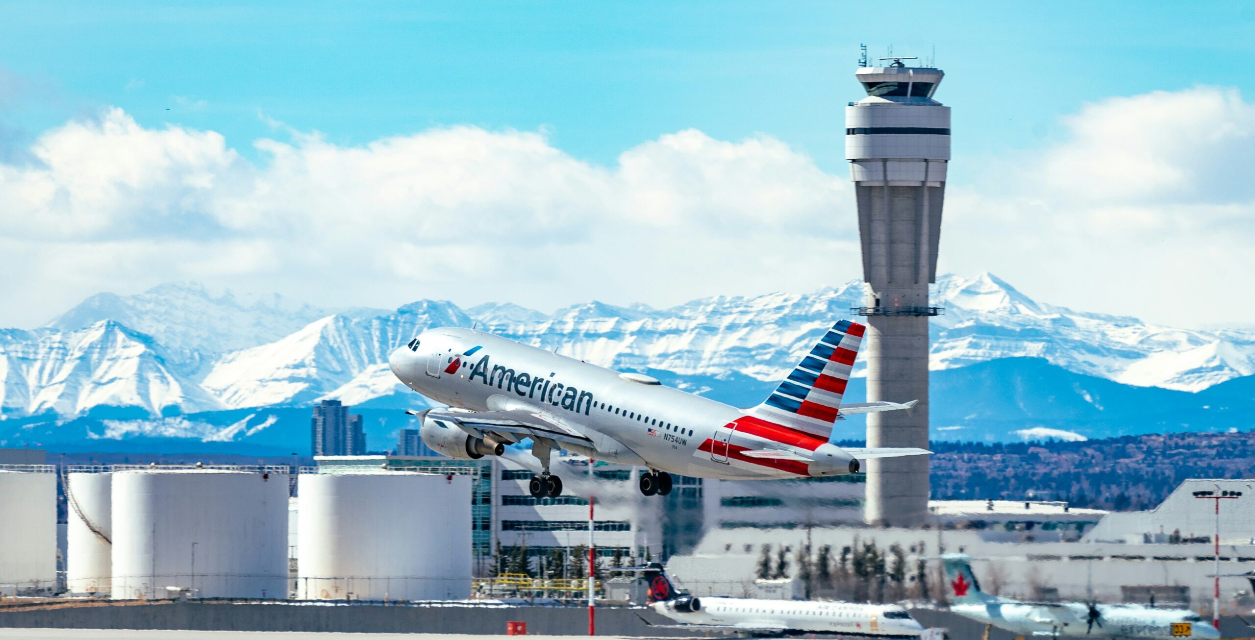 An American Airlines passenger jet takes off from an airport runway, climbing beside a tall air traffic control tower. Snow-covered mountains stretch across the background under a bright blue sky with scattered clouds, while airport fuel tanks, terminal buildings, and an Air Canada aircraft are visible in the foreground