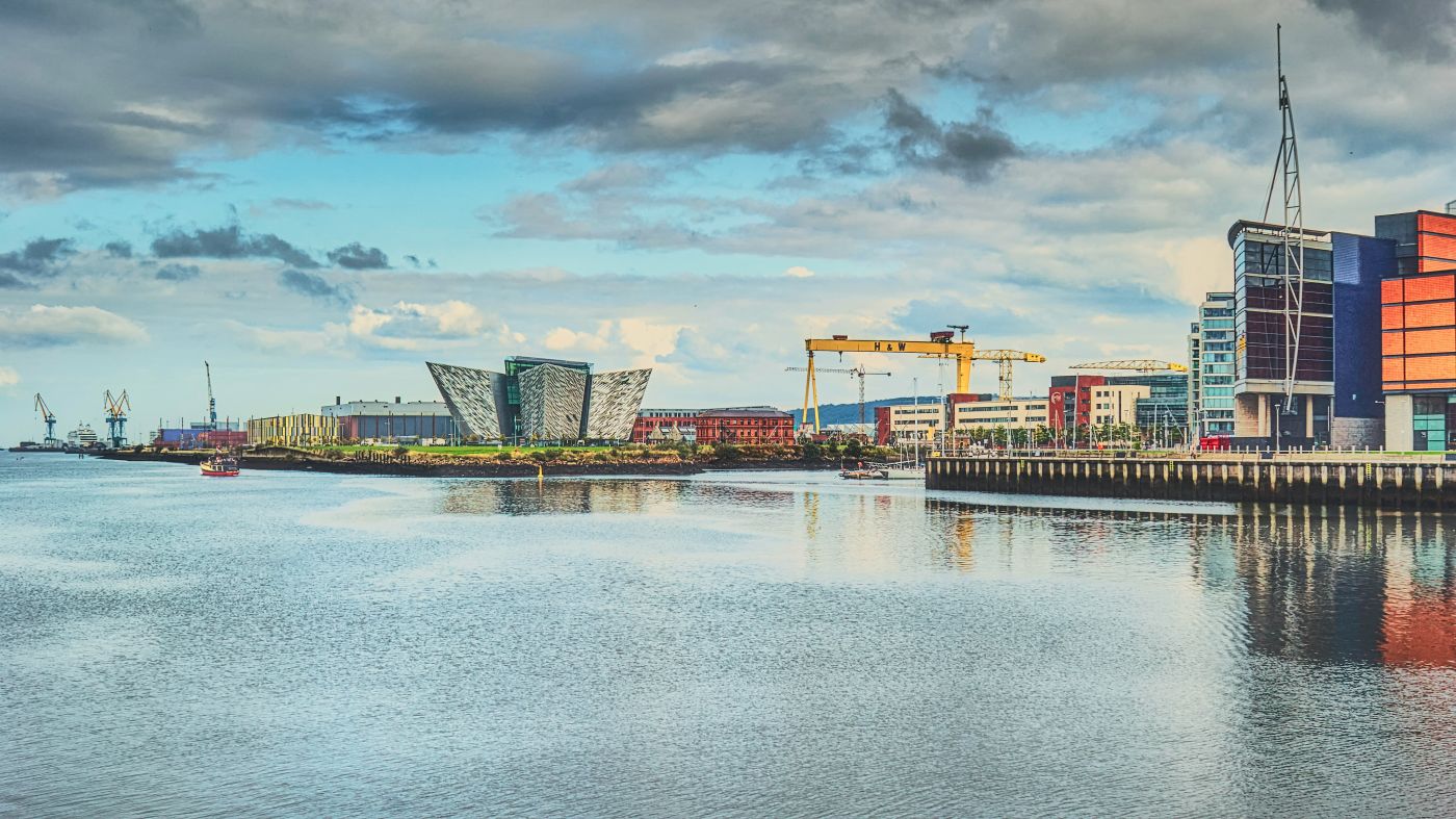 A cityline view of belfast from the water, with the River Lagan's crystal water in the front f the image and various high rise towers and port equipment seen in the horizon