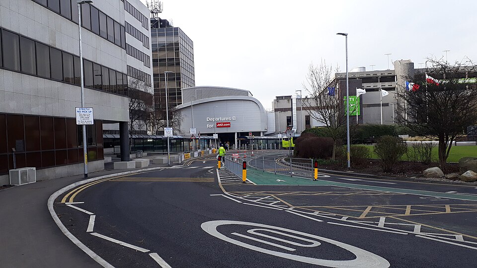 Exterior of Manchester Airport with the sign 'Departures B' above the entrance. The road leading to the terminal can be seen in the front of the photo.