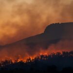 An image of a large wildfire in a forest with the silhouette of a mountain or large hill in the background. The sky in the background is orange and filled with smoke from the blaze.