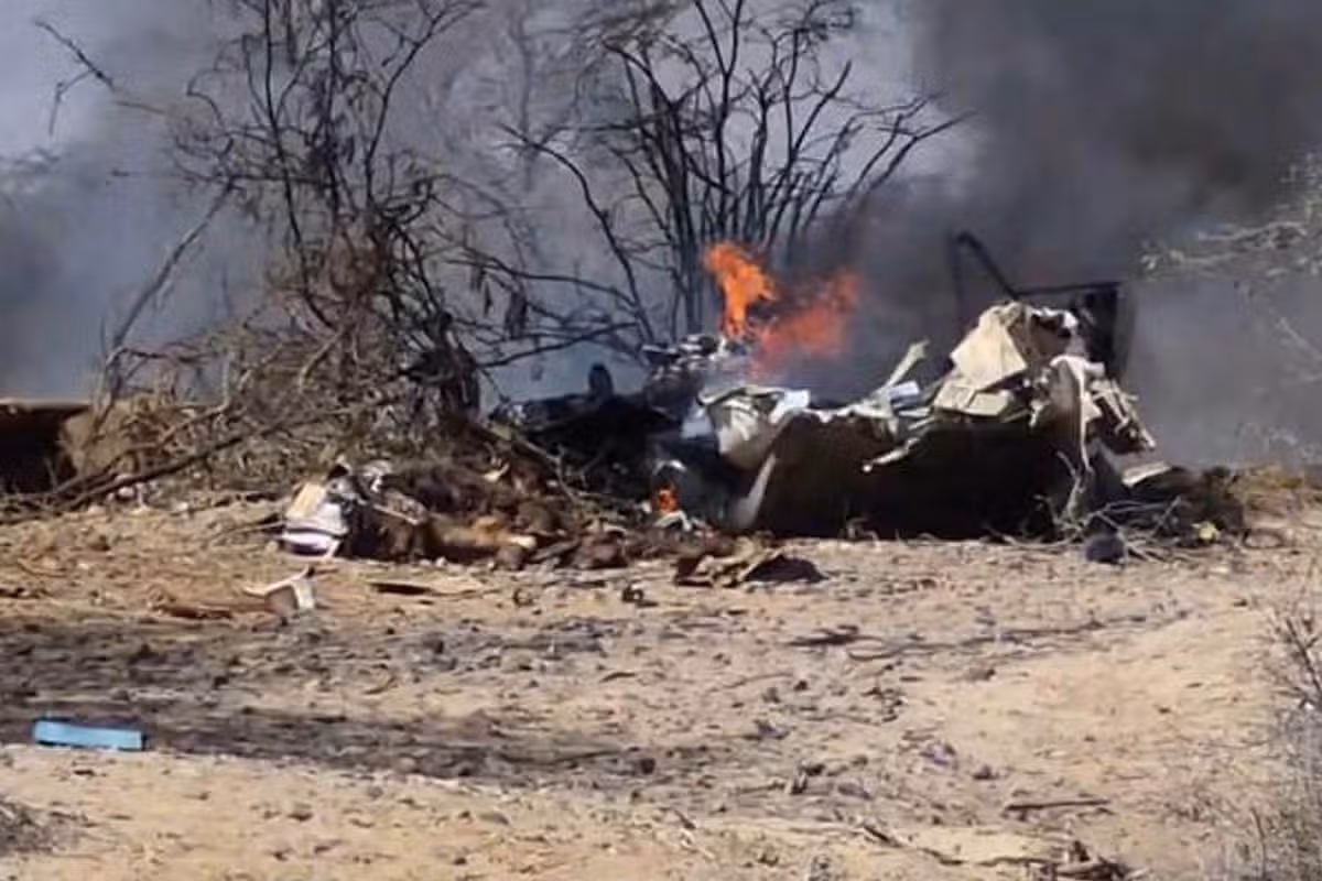The wreckage of the plane crash can be seen with the parts of the plane scattered, and some on fire. Burnt and knocked down trees can be seen through the smoke behind the wreckage.