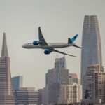United Airlines wide-body passenger jet flying across the San Francisco skyline, framed between the Transamerica Pyramid and Salesforce Towe