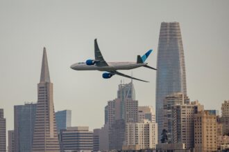 United Airlines wide-body passenger jet flying across the San Francisco skyline, framed between the Transamerica Pyramid and Salesforce Towe