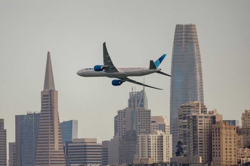 United Airlines wide-body passenger jet flying across the San Francisco skyline, framed between the Transamerica Pyramid and Salesforce Towe