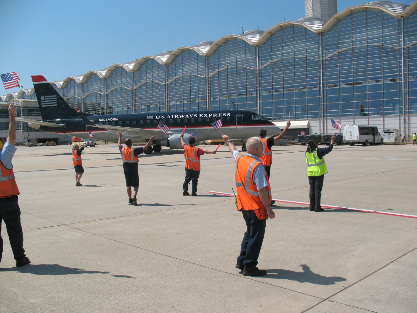 Ronald Reagan Washington National Airport tarmac with employees in high-visibility jackets waving US flags, and a US Airways Express aircraft behind them, the background has a view of a clear blue sky and an airport terminal