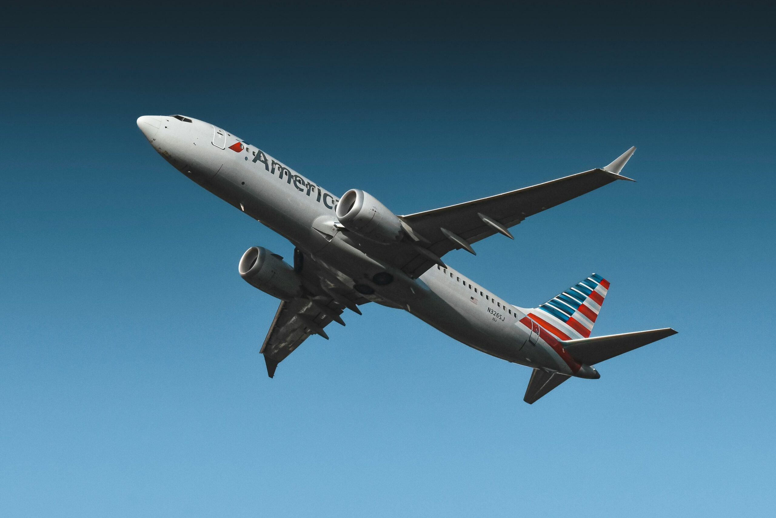 Low-angle view of an American Airlines passenger jet in flight against a clear blue sky. The aircraft’s silver fuselage, twin engines, and red, white, and blue striped tail are clearly visible as it climbs overhead with landing gear retracted