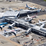 An overview shot of Sydney Airport (SYD) showing the exterior of the airport building with planes parked on the tarmac surrounding the hub. Lots of smaller vehicles can also be seen.