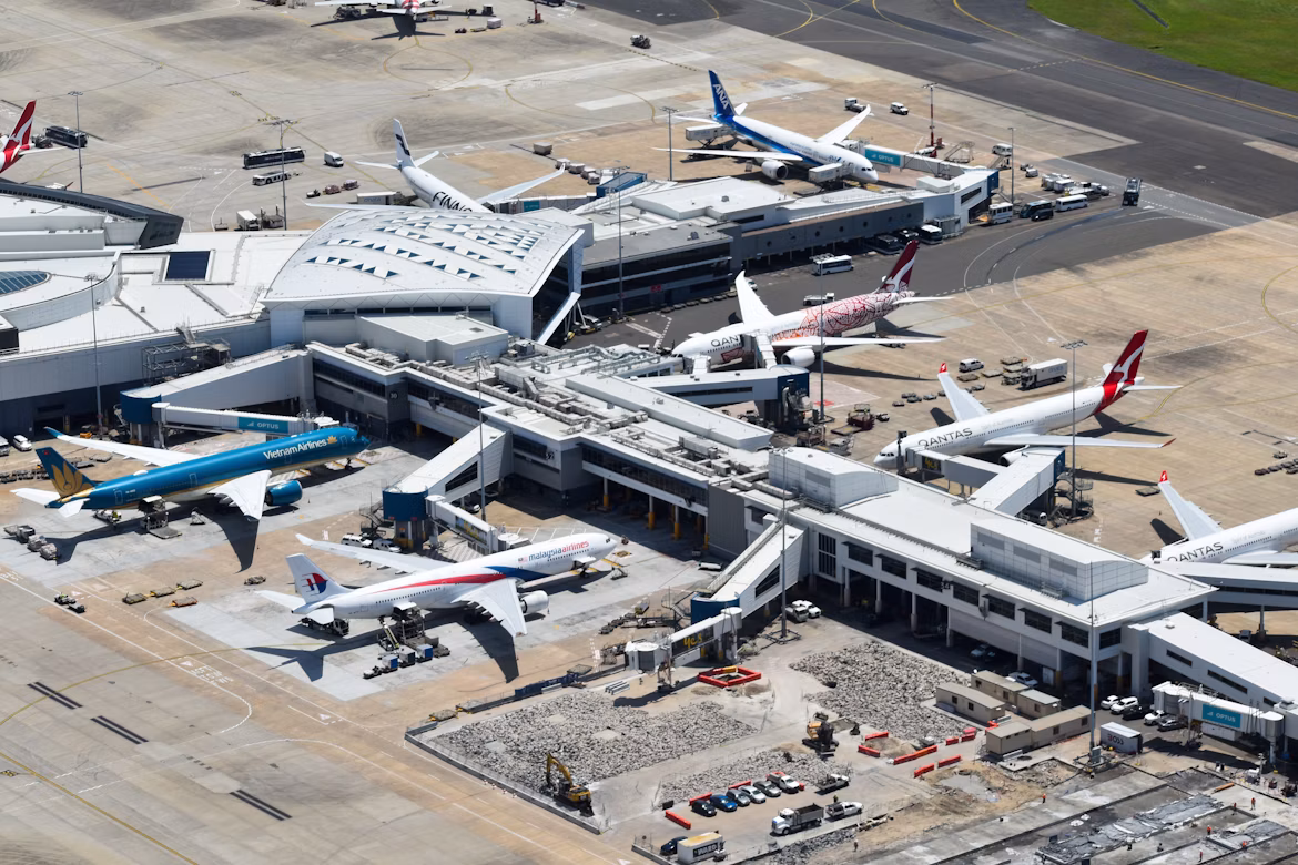 An overview shot of Sydney Airport (SYD) showing the exterior of the airport building with planes parked on the tarmac surrounding the hub. Lots of smaller vehicles can also be seen.