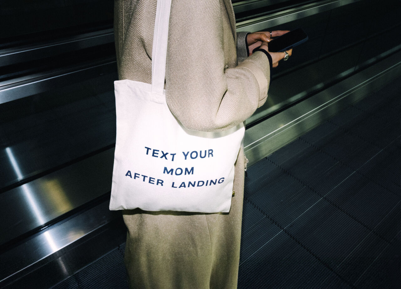 A faceless person stands on an escalator, wearing a tote bag that reads