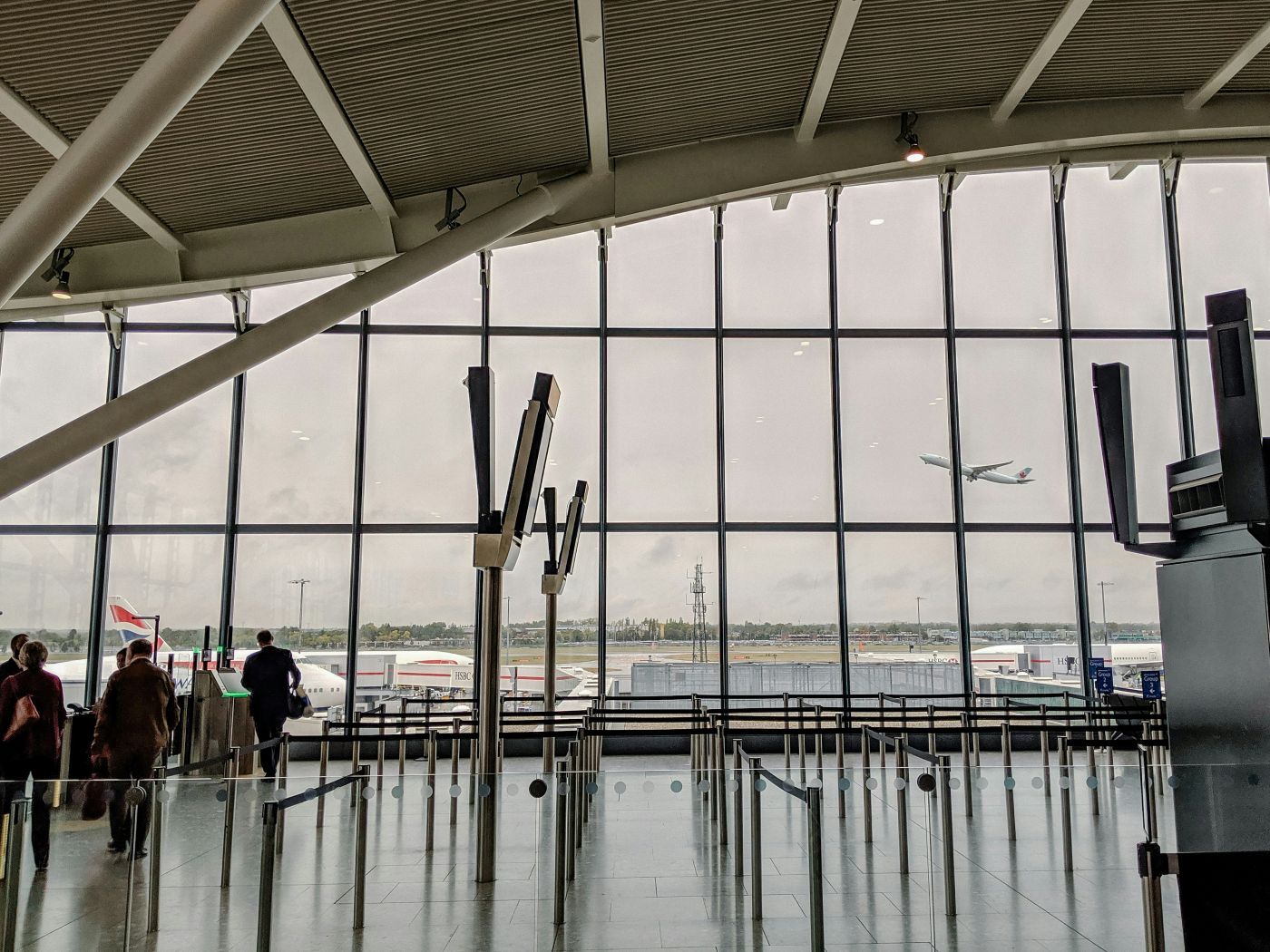 A view of a Heathrow terminal showing a large see through glass wall with the clear sky and an aircraft ascending in the horizon, people are walking in the terminal