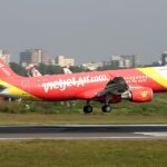 A VietJetAir A320 midflight above a concrete runway with buildings being able to be seen just behind the aircraft.