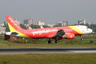 A VietJetAir A320 midflight above a concrete runway with buildings being able to be seen just behind the aircraft.