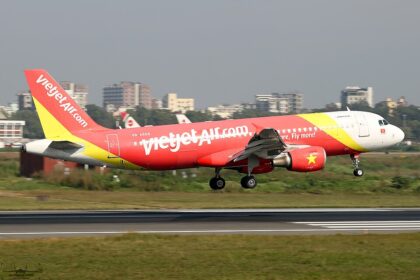 A VietJetAir A320 midflight above a concrete runway with buildings being able to be seen just behind the aircraft.