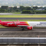 A VietJetAir A321 plane sat on a concrete runway with green grass and foliage in the background.