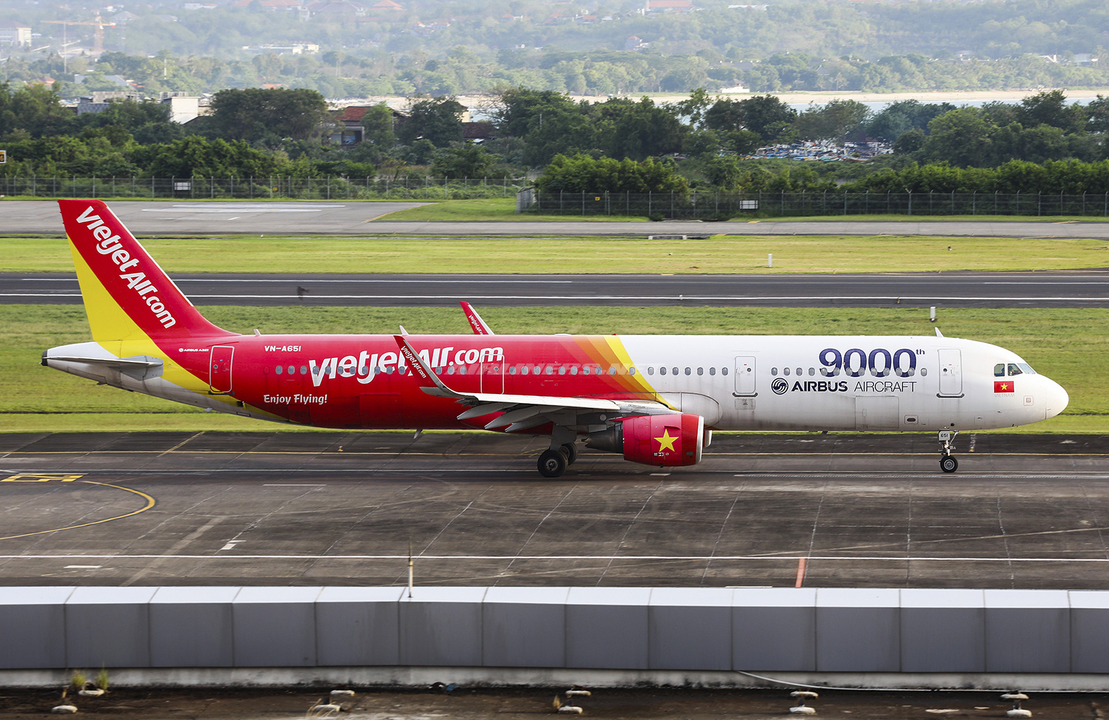 A VietJetAir A321 plane sat on a concrete runway with green grass and foliage in the background.