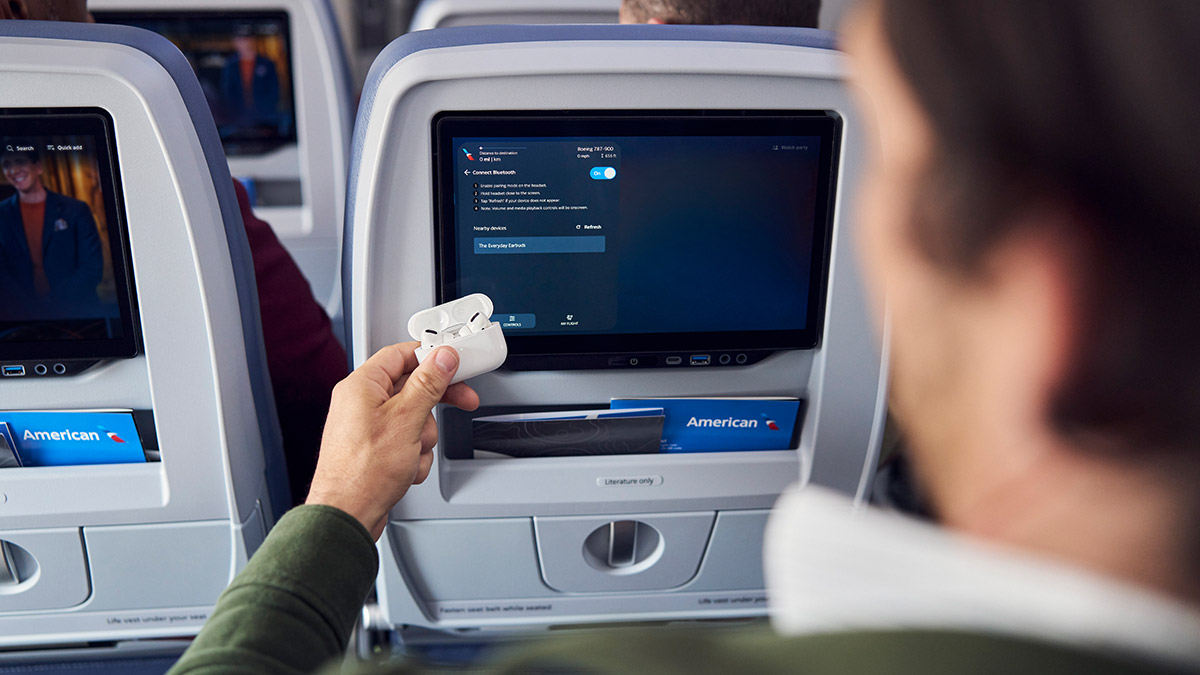 An airline passenger holding an open white earbud case up to a seatback entertainment screen showing a