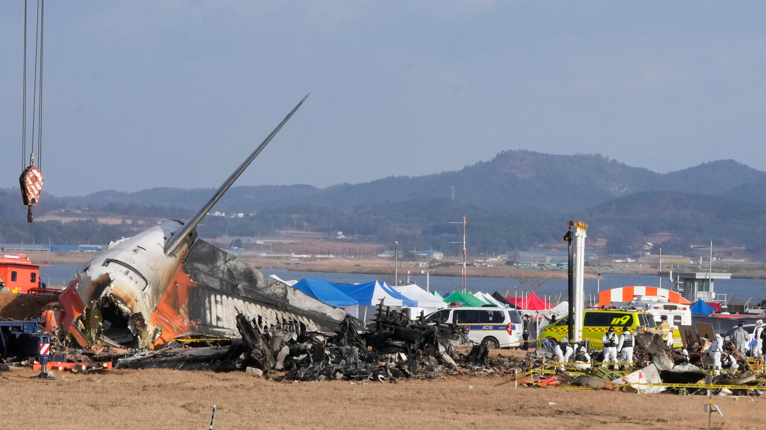 An image from the aftermath of the Jeju Air Crash on December 29 2024. The burnt remains of the backend of the Boeing 737-800 an be seen on the left, with further blackened aircraft ruins in the centre and background. People in hazmat suits investigate the ruins. Mountains and a shoreline be seen in the background through foggy air. 