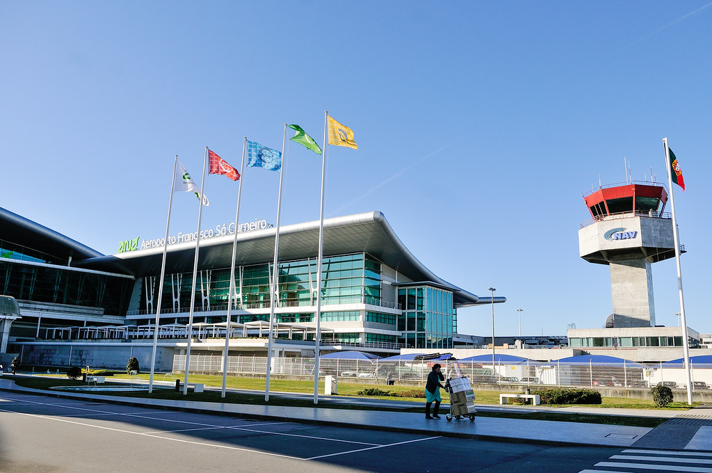 Exterior view of Francisco Sá Carneiro Airport, with the glass terminal, waving flags and the control tower in the background.