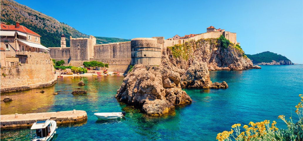 Image shows the Dubrovnik in Croatia with castle walls on top of cliffs, boats visible on clear sea water with clear blue skies