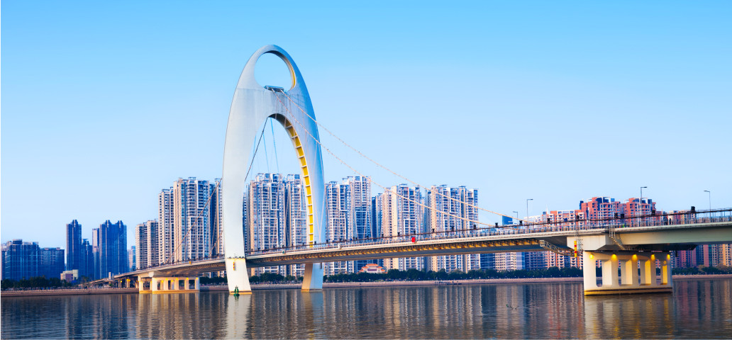Image shows a futuristic bridge over the Pearl River Delta with skyscrapers in the background on a clear day in Guangzhou