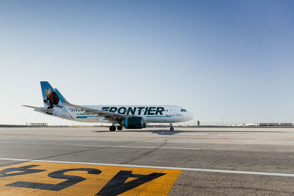 A white Frontier Airlines passenger plane with the green logo on the fuselage and the image of a red panda on the tail, parked in broad daylight on the concrete runway of an airport under a clear blue sky.
