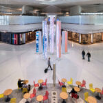 Elevated view of Newark Liberty International Airport terminal A with a central digital art installation of tall, glowing pillars. High-end retail stores line the concourse. Travelers walk through the space, and in the foreground, people sit at tables with red and yellow chairs.