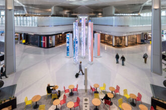Elevated view of Newark Liberty International Airport terminal A with a central digital art installation of tall, glowing pillars. High-end retail stores line the concourse. Travelers walk through the space, and in the foreground, people sit at tables with red and yellow chairs.