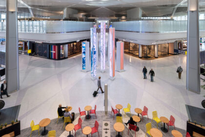 Elevated view of Newark Liberty International Airport terminal A with a central digital art installation of tall, glowing pillars. High-end retail stores line the concourse. Travelers walk through the space, and in the foreground, people sit at tables with red and yellow chairs.