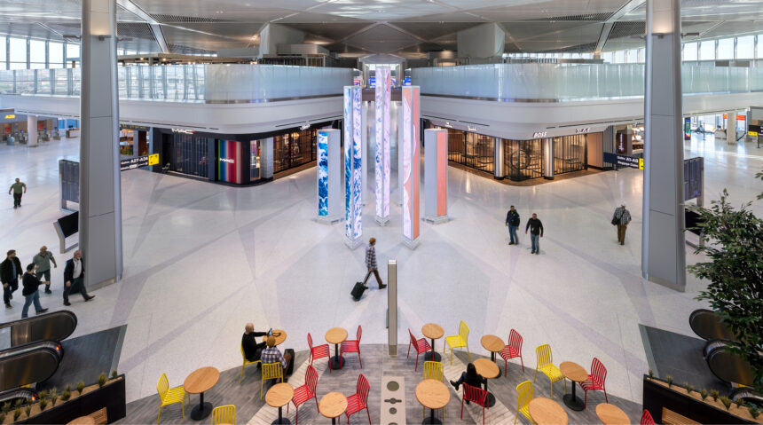 Elevated view of Newark Liberty International Airport terminal A with a central digital art installation of tall, glowing pillars. High-end retail stores line the concourse. Travelers walk through the space, and in the foreground, people sit at tables with red and yellow chairs.