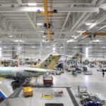 Wide high-angle view inside a bright, vast airplane manufacturing hangar showing multiple commercial aircraft undergoing assembly.