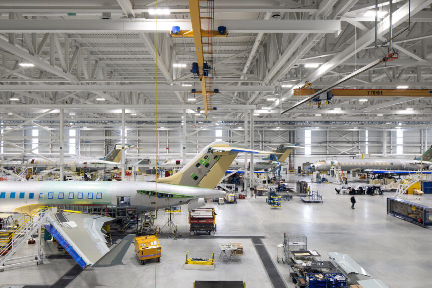 Wide high-angle view inside a bright, vast airplane manufacturing hangar showing multiple commercial aircraft undergoing assembly.