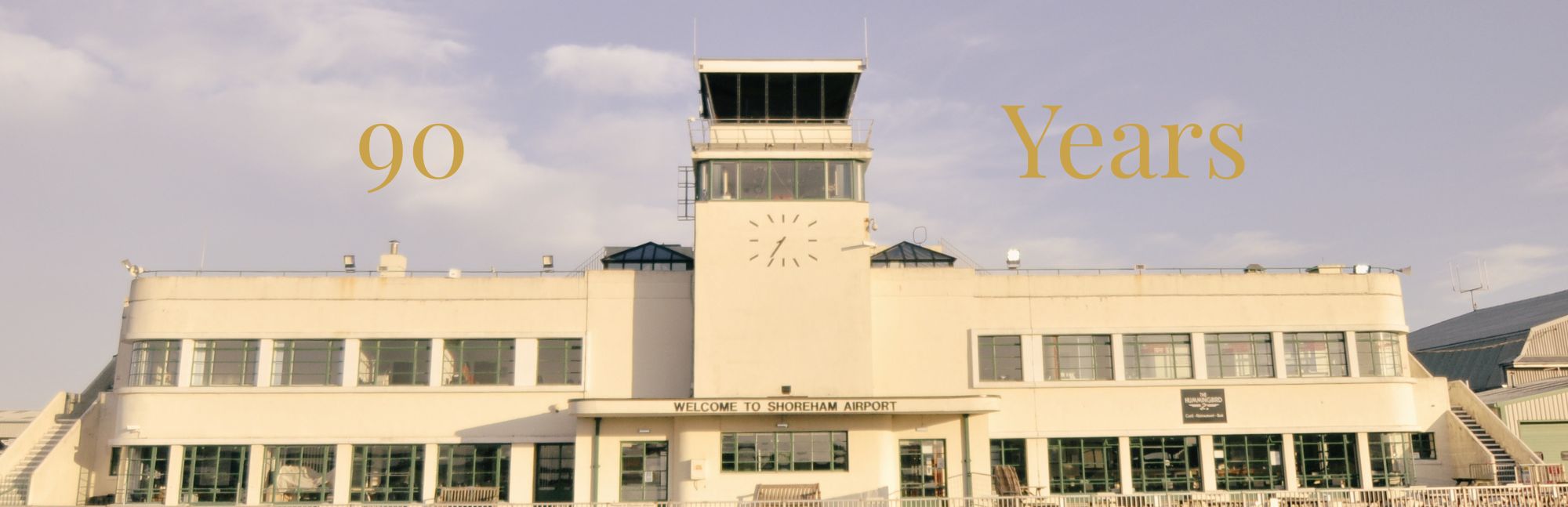 Image shows the Art-Deco 1936 Brighton City Airport terminal building in Lancing/Shoreham, West Sussex on a cloudy day with 90 years written in the sky