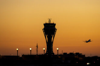 The outline of an Air Traffic Control tower can be seen in front of a sunset illuminated sky. The outline of an aircraft is seen flying away from the tower.