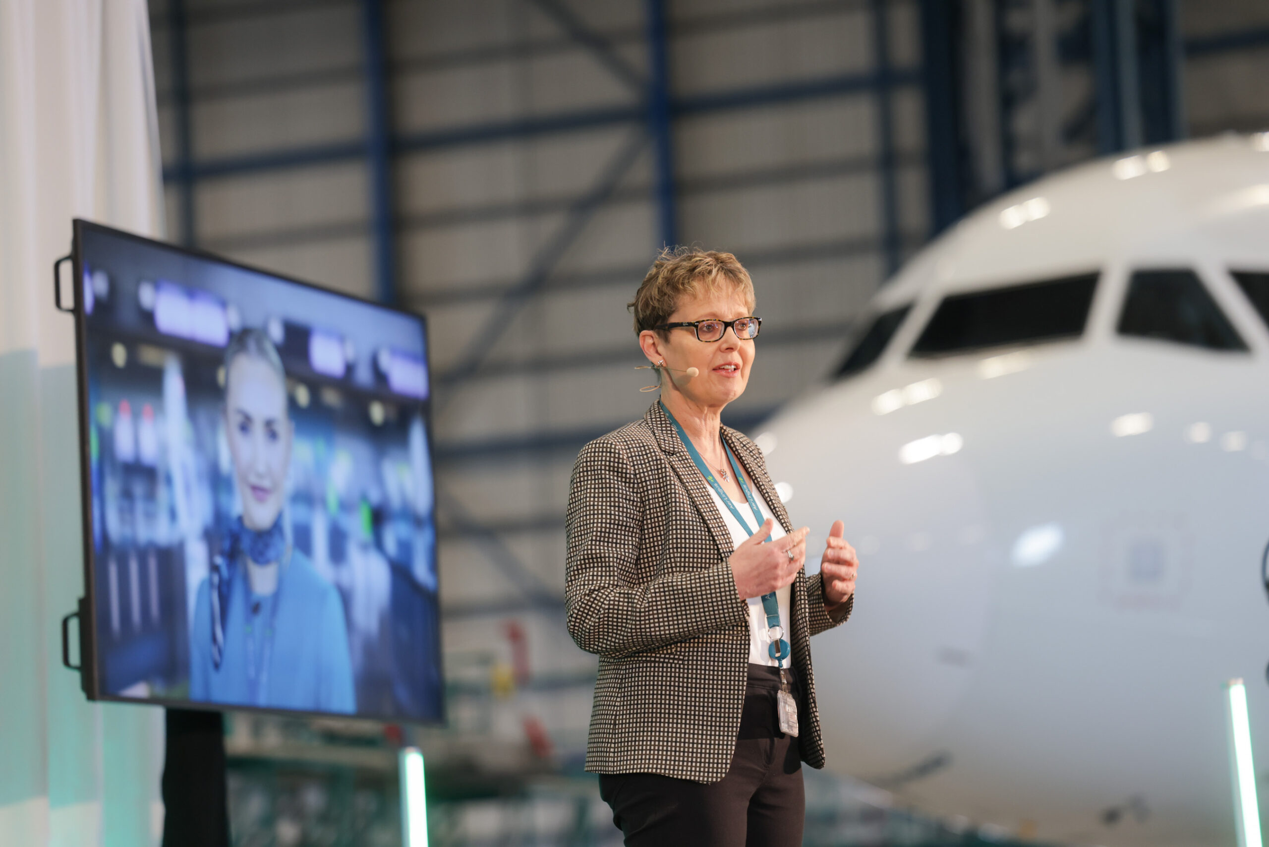 Image shows the CEO of airline Aer Lingus, Lynne Embleton in an aircraft hangar giving a presentation next to an aircraft and screen