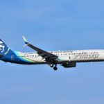 An Alaskan Airlines Boring 737 MAX 9 is pictured in flight. It is mostly white with a dark blue tail that has the airline's logo of a stylised native's face on it. The nose is positioned on the far right, the tail far left. A bright, clear blue sky with no clouds occupies the background