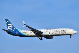 An Alaskan Airlines Boring 737 MAX 9 is pictured in flight. It is mostly white with a dark blue tail that has the airline's logo of a stylised native's face on it. The nose is positioned on the far right, the tail far left. A bright, clear blue sky with no clouds occupies the background