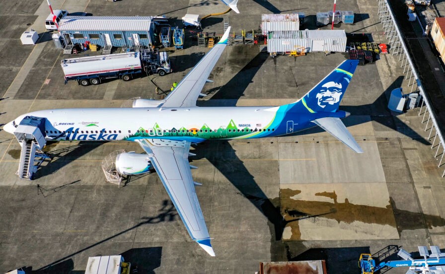 A Boeing 737 MAX 9 is pictured on the ground from above. It is an Alaskan Airlines aircraft, so it is mostly white, but has a blue tail with the logo of a stylised Alaskan Native person's face printed in white, and green trees printed along the lower fuselage. The aircraft is an ecoDemonstrator during engine emissions testing, positioned in the centre of the picture, nose on the far left and tail on the far right. The background depicts concrete ground, with metal storage containers and vehicles surrounding the plane.