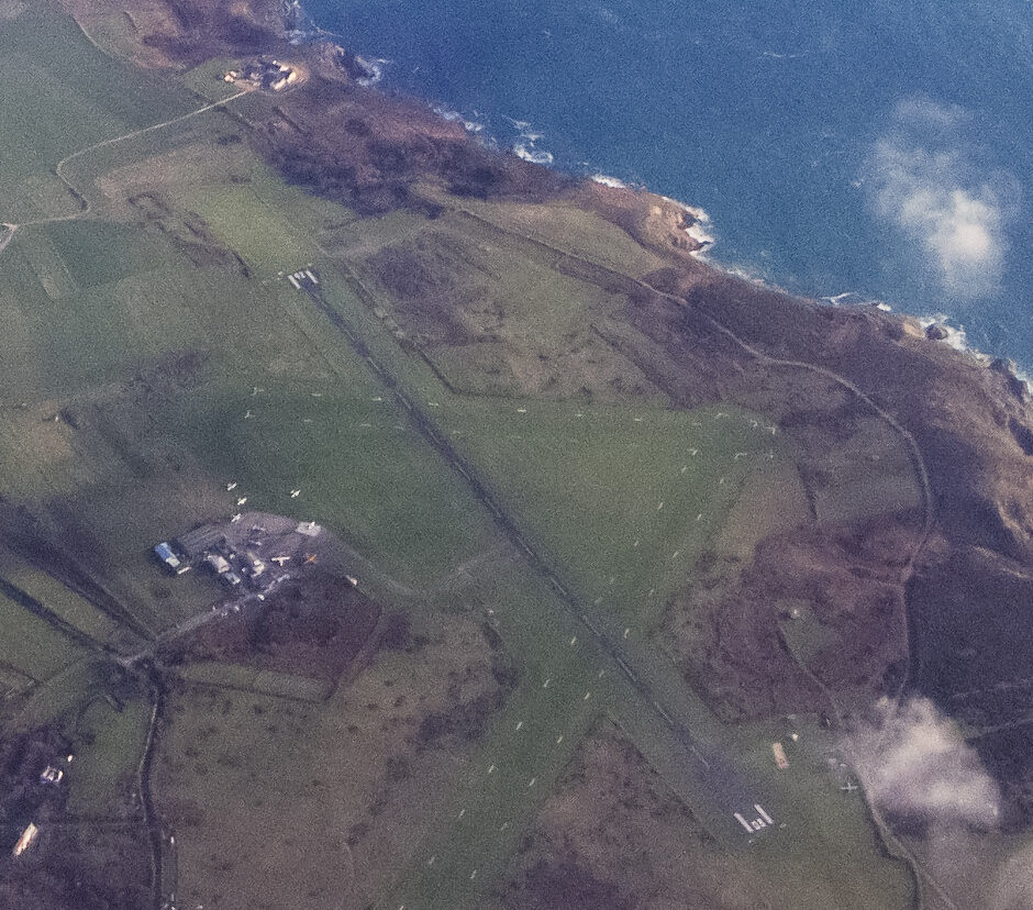 Aerial view of Alderney Airport. In the centre, the runway runs diagonally from upper left to lower right, with the airport buildings to the lower let. Grass fields surround the runway and airport, and hills ascend on the lower far right side of the image. In the top right of the image, the shorelines can be seen, with blue sea evident below some cloudiness.